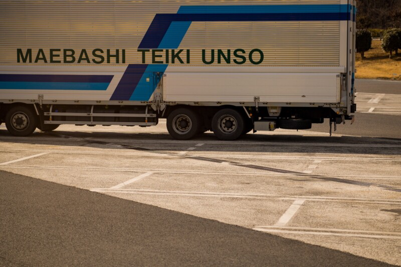 A white and blue truck with "MAEBASHI TEIKI UNSO" on the side, parked on an asphalt surface. The landscape around is dry with sparse vegetation, and the truck occupies most of the frame.