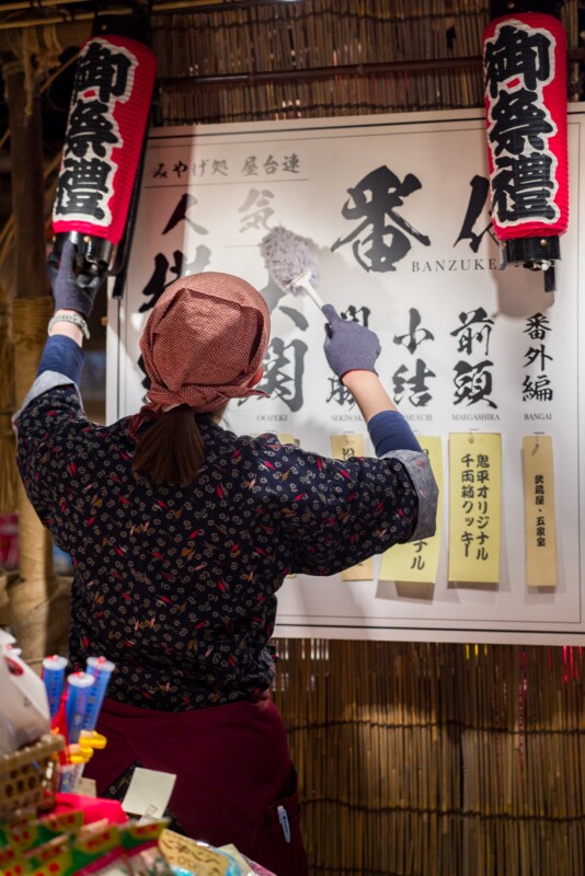 A person wearing a patterned shirt, headscarf, and gloves is cleaning a large sign or poster with Japanese characters, under decorative red lanterns. Various snacks or goods are visible in the foreground.