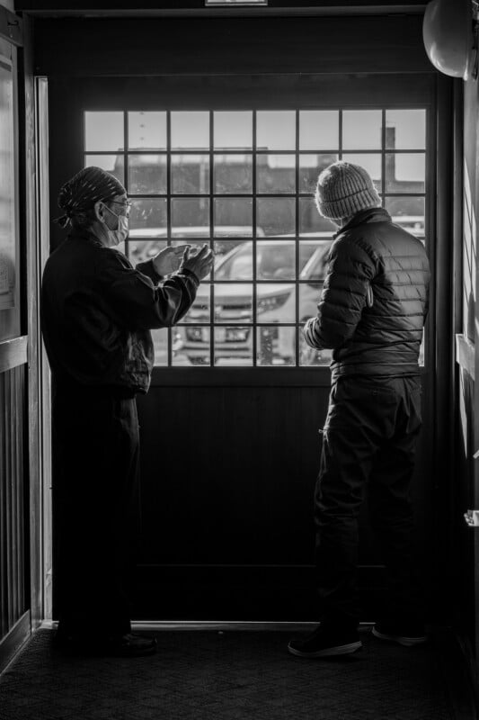 Two people standing by a glass-paneled door, engaged in conversation. One person is wearing a hat and gesturing with hands, while the other wears a beanie and looks outside. A row of parked cars is visible through the door. Black and white image.