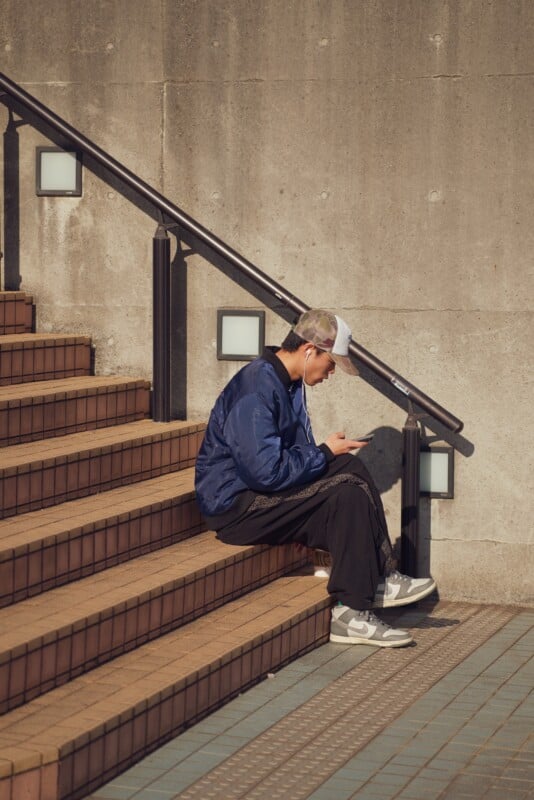 A person wearing a blue jacket, black pants, and a cap sits on outdoor stairs. They are looking at their phone with earphones in. The stairs have tiled surfaces and a metal railing. The scene is well-lit with late afternoon sunlight.
