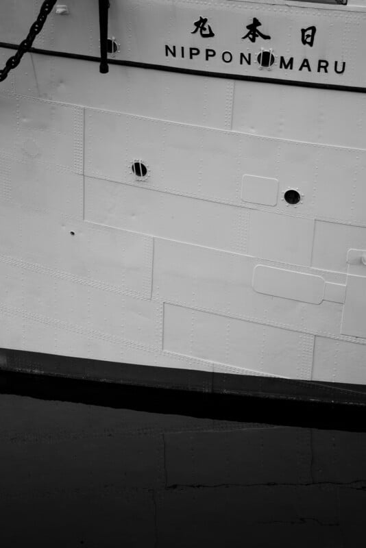 Close-up of a ship's hull in black and white, featuring the name "Nippon Maru" and Japanese characters above. The surface has visible panels and rivets, with a portion of the waterline and a chain visible on the left side.