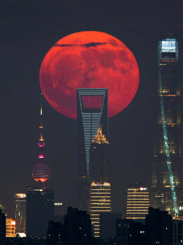 A large, red full moon appears to rise directly behind modern skyscrapers in a city skyline at night, including the Shanghai World Financial Center and the Oriental Pearl Tower, creating a dramatic and vivid urban scene.