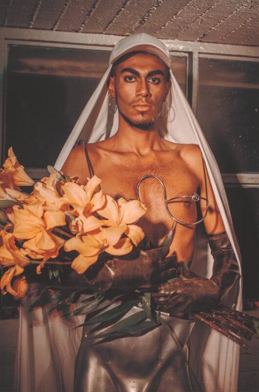 A person wearing a white veil and cap poses indoors, holding a bouquet of orange lilies. They wear metallic gloves and a unique, futuristic outfit with hoop earrings and dramatic makeup.