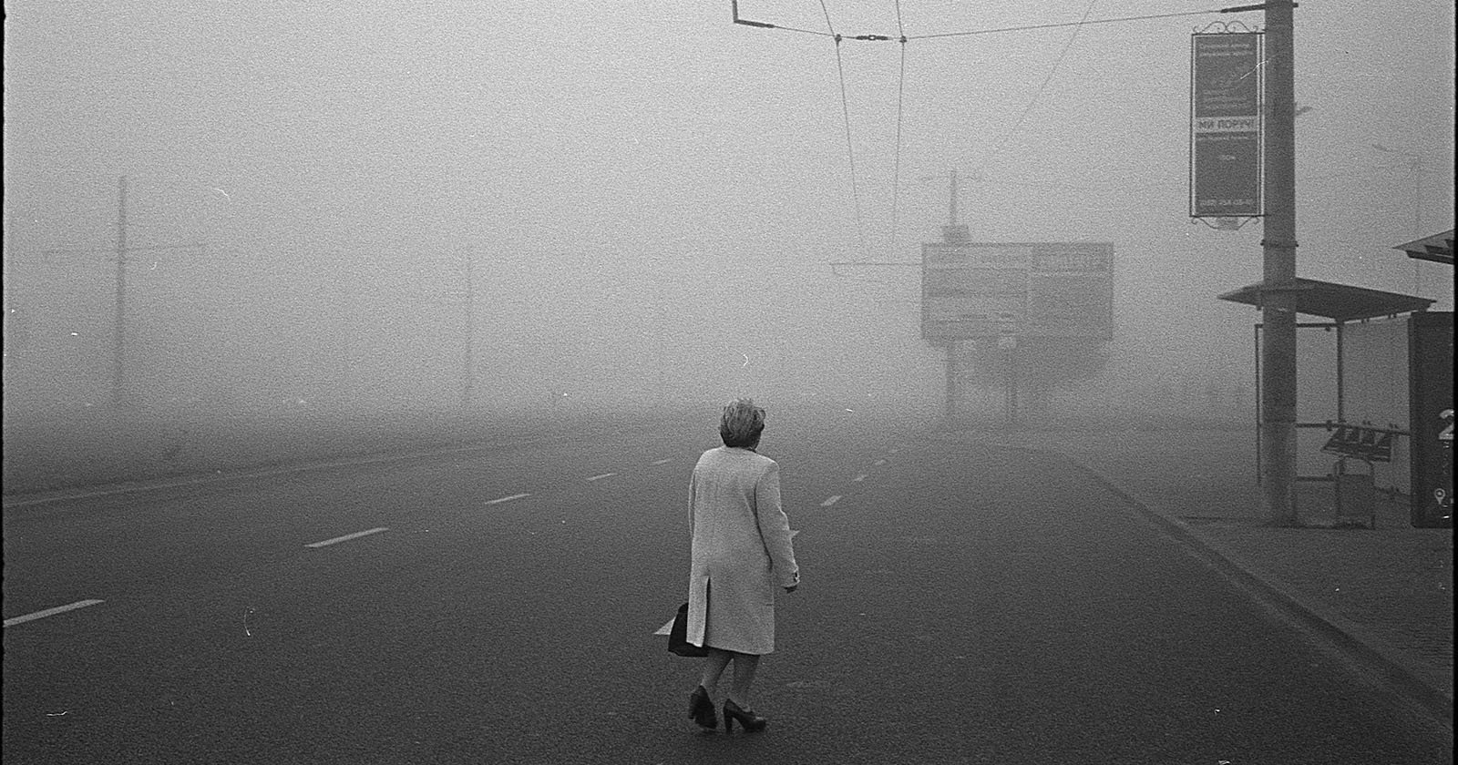 A person in a light coat walks alone on an empty, foggy street. The surroundings are barely visible due to thick fog, with street signs and electric poles fading into the background.