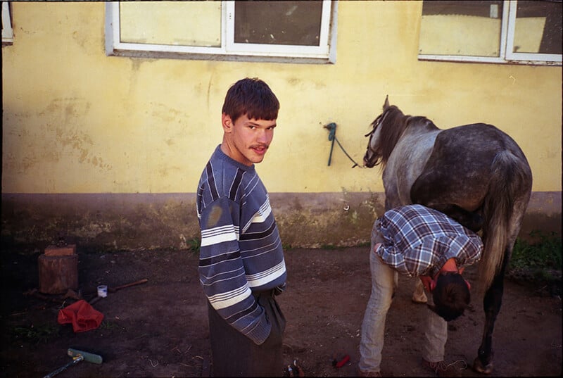 A man in a striped sweater stands looking at the camera, while another person bends down tending to a gray horse’s hoof. They are outside near a yellow wall with windows. Tools and objects are scattered on the ground.