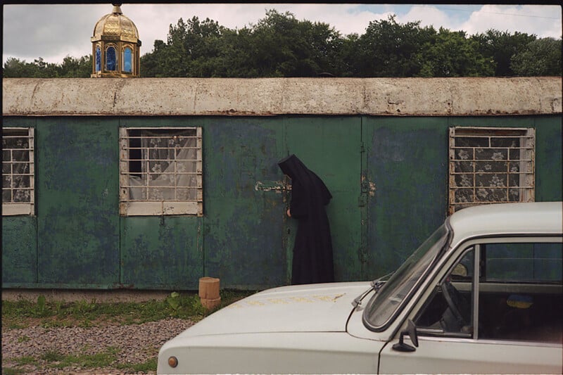 A person in a black robe unlocks a green, weathered metal building with barred windows. A white car is parked in front, and a golden dome rises above the trees in the background.