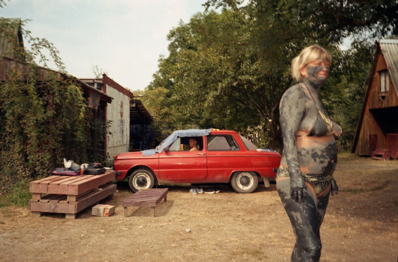 A woman covered in dark mud stands outside near a red vintage car; a person is visible inside the car. Wooden benches and trees are in the background on a sunny day.