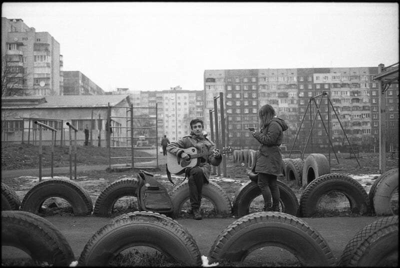 A person sits on a tire playing a guitar while another stands nearby looking at a phone. They are in an urban playground with large tires, swings, and apartment buildings in the background on a cloudy day.