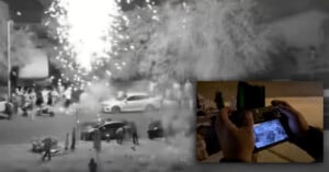 A nighttime street scene shows fireworks exploding near a crowd and parked cars. Inset at the lower right, hands hold a controller, viewing a live camera feed of the event.