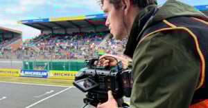 A cameraman in a green jacket films a motorsport event at a racetrack, with grandstands full of spectators and Michelin banners visible in the background.