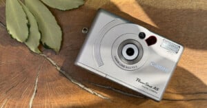 A silver Canon PowerShot A5 digital camera rests on a wooden surface near some green and yellow leaves, with sunlight casting shadows across the scene.