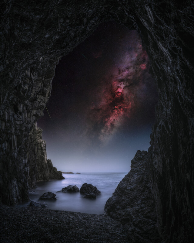View from inside a dark rocky cave looking out to a calm sea under a dramatic night sky, featuring a vivid, colorful galaxy and stars illuminating the scene beyond the cave entrance.