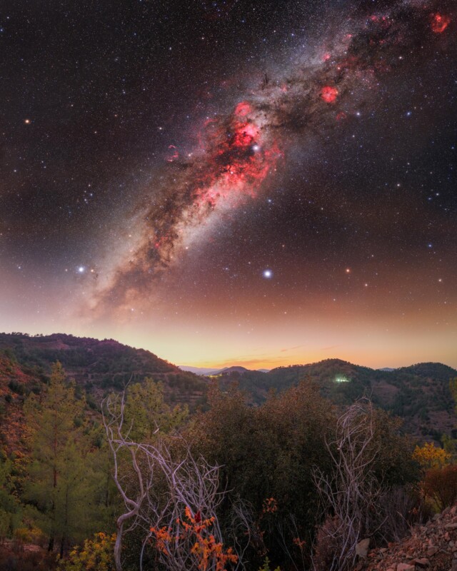 A vibrant Milky Way galaxy stretches across a starry night sky above pine-covered hills and a valley, with dry bushes and trees in the foreground under a fading sunset.