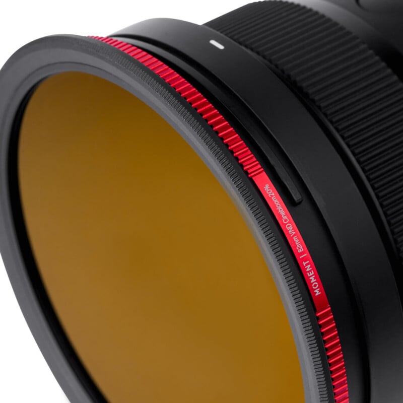 Close-up of a camera lens with a circular amber-colored filter attached. The filter has a red textured rim with the text "MOMENT 82mm VND Cinefilter" engraved on it.