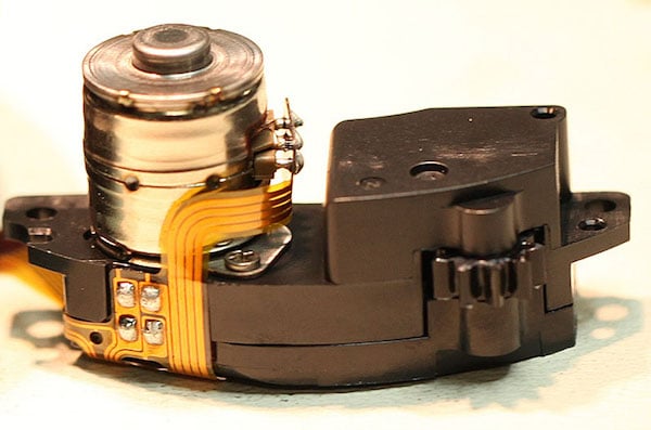 Close-up of a hard disk drive (HDD) head actuator assembly with a visible metal motor, orange flex cable, and black plastic components against a light background.