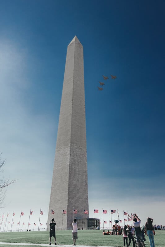 People watch and photograph a formation of four jets flying over the Washington Monument on a clear day, with American flags surrounding the monument and a blue sky in the background.
