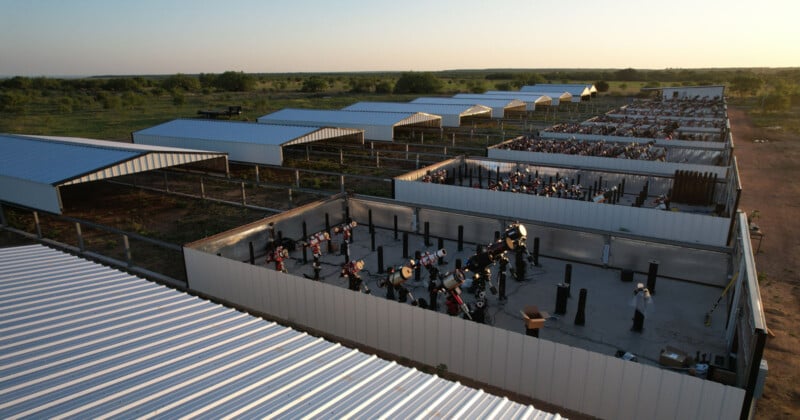 Aerial view of multiple fenced enclosures with dozens of drum sets and percussion instruments arranged in rows, covered by metal roofs, set in an open rural landscape at sunset.