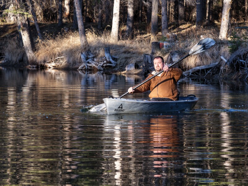 A man in a brown jacket paddles a gray kayak on a calm lake, surrounded by trees and dry grass along the shoreline, with sunlight casting reflections on the water.