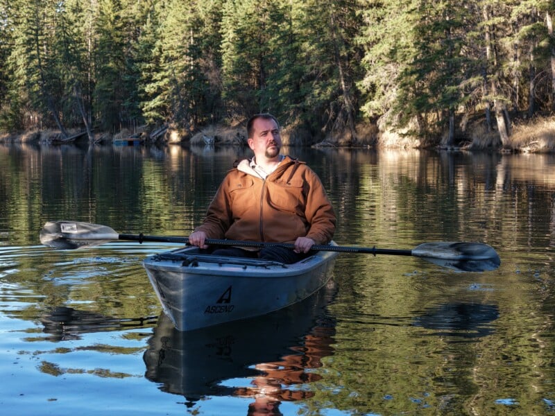 A man in a brown jacket sits in a kayak on a calm lake, holding a paddle. He is surrounded by tall evergreen trees, and the water reflects the forest and sky.