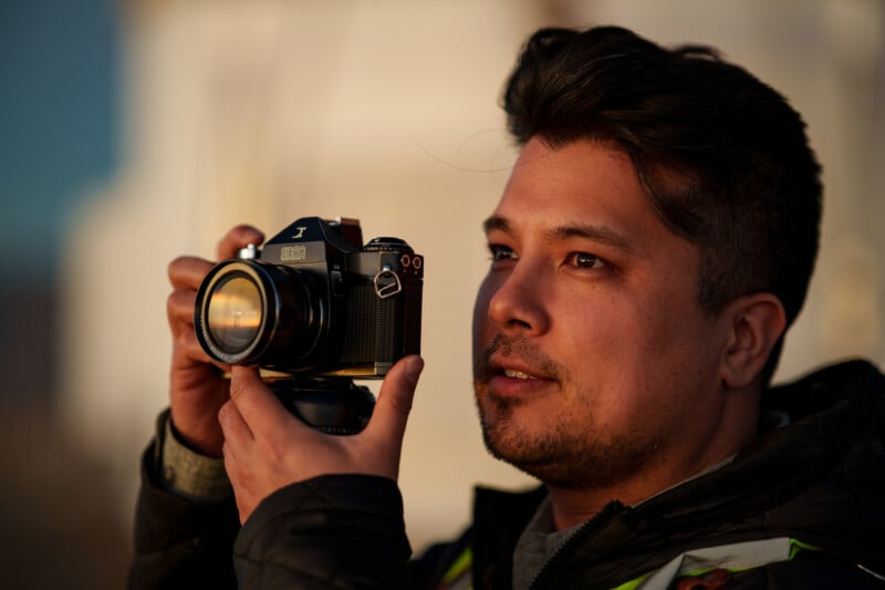 A man with dark hair and facial hair holds a vintage camera up to his face, looking attentively at his subject. The lighting is warm and soft, suggesting sunset or sunrise.