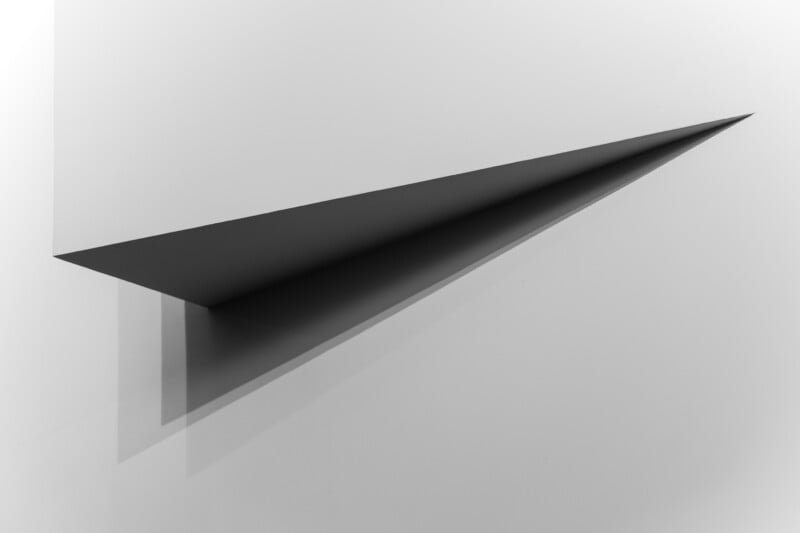 A minimalist black sculpture resembling a paper airplane juts from a white wall, casting a sharp triangular shadow that accentuates its geometric shape.