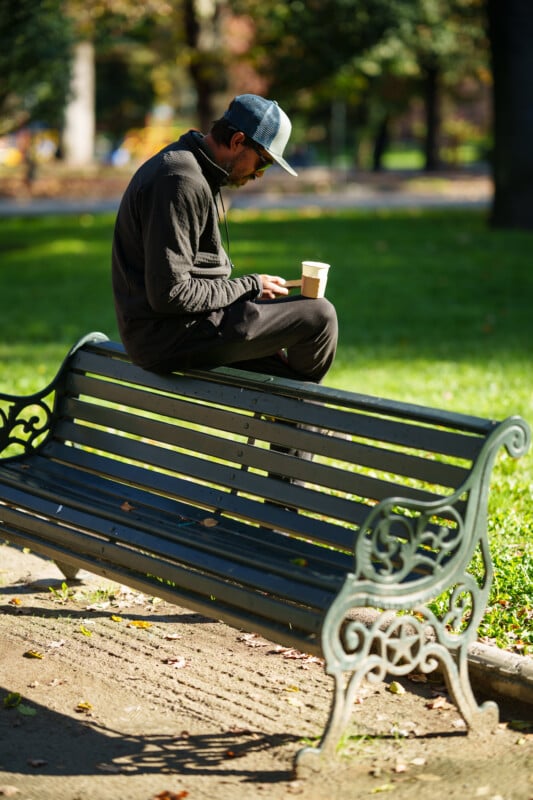 A man wearing a cap and dark clothing sits cross-legged on top of a park bench, looking at his phone and holding a coffee cup, with green grass and trees in the background.