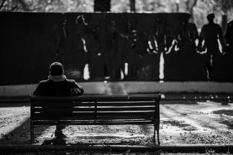 A person sits alone on a park bench in black and white, facing a wall with cut-out human figures, surrounded by scattered leaves and soft sunlight.