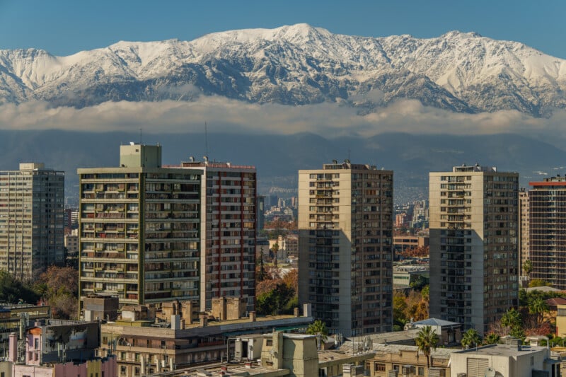 Tall apartment buildings rise before distant snow-capped mountains under a clear blue sky, with a layer of clouds partially covering the mountain slopes.