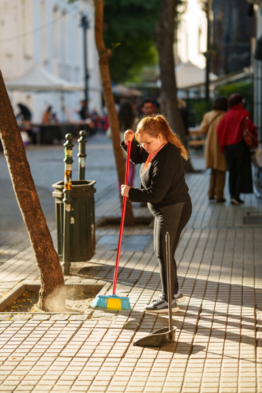 A woman in black clothing sweeps a city sidewalk with a broom and dustpan on a sunny day; trees, a trash bin, and people are visible in the background.