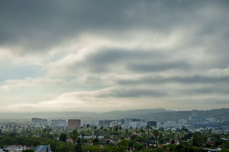 A cityscape under a cloudy sky, with low, thick clouds and morning haze. Tall buildings and palm trees are visible, surrounded by greenery and houses, with distant hills in the background.