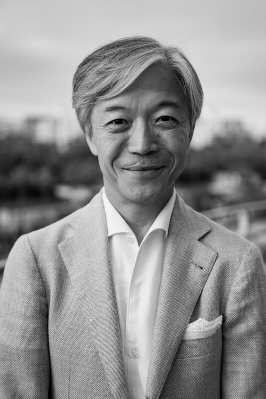 A man with gray hair wearing a light-colored suit jacket and a white shirt smiles at the camera outdoors, with blurred trees and buildings in the background. The image is in black and white.