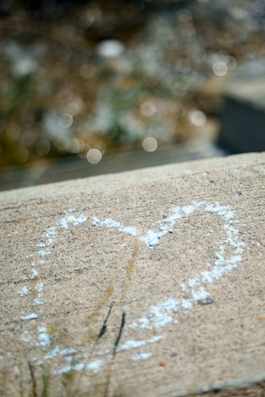 A pale blue heart is drawn with chalk on a rough concrete surface, with a blurred natural background in soft, warm light.
