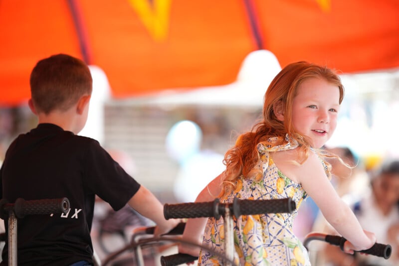 A young girl with red hair in a patterned dress smiles while riding a carousel, with a boy in a black shirt beside her. The bright orange canopy is visible overhead.