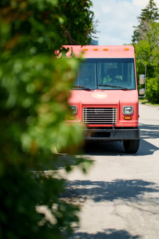 A red delivery truck is parked on a sunlit street, partially obscured by leafy green branches in the foreground. The vehicle's front is visible, and the driver can be seen through the windshield.