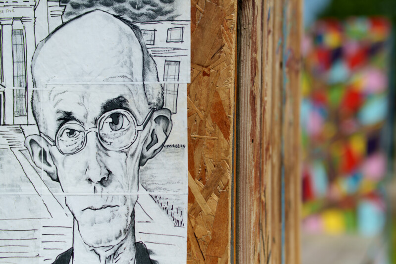 A black and white drawing of a serious, bald man with glasses is posted on a wooden wall. The background is blurred and colorful, contrasting with the monochrome sketch.