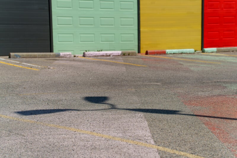 A parking lot with the shadow of a streetlamp on the pavement. In the background are garage doors painted in green, yellow, and red. The ground has faint lines and pastel-colored markings.