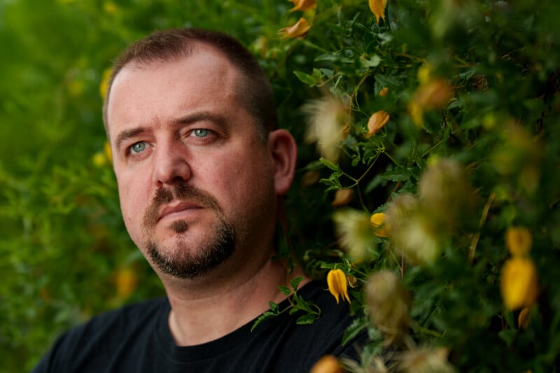 A man with short hair and a beard stands in front of green foliage with yellow flowers, gazing thoughtfully into the distance. The background is softly blurred, highlighting his face.