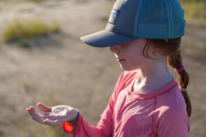A young girl in a pink shirt and gray cap looks at something small resting on her open palm while standing outdoors in sunlight.