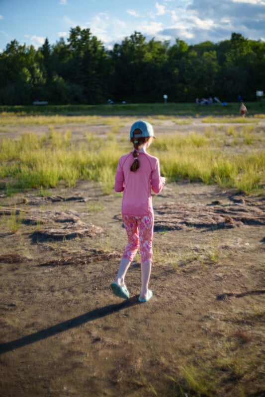 A young girl wearing a blue cap, pink long-sleeve shirt, and pink leggings walks outdoors on a grassy, sunlit field with trees in the background.