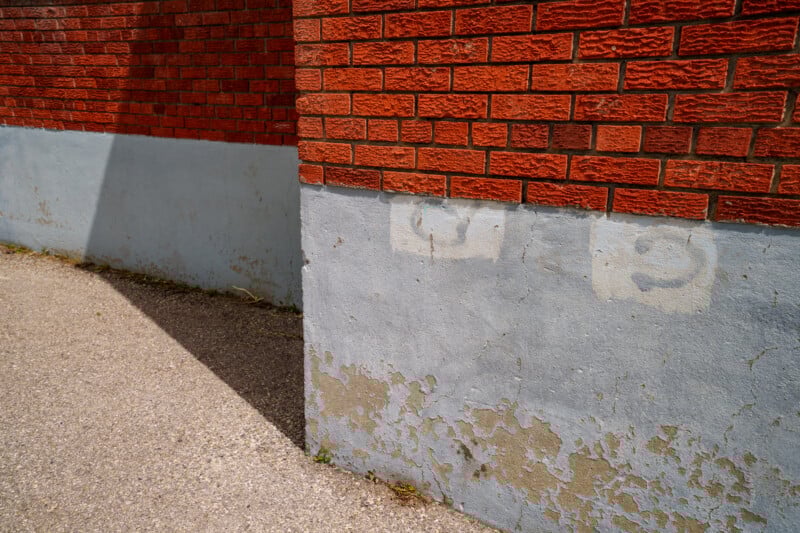 A red brick wall with a peeling, light blue painted lower section meets a paved ground in bright sunlight, creating sharp shadows and visible weathered textures on the building’s surface.