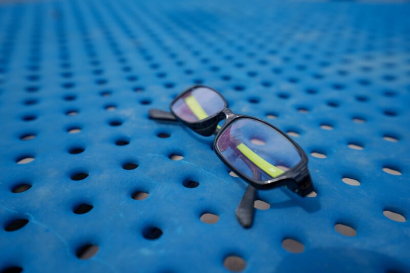 A pair of black-rimmed eyeglasses rests on a blue perforated metal surface, with the glasses’ arms open and the surface extending into the blurred background.