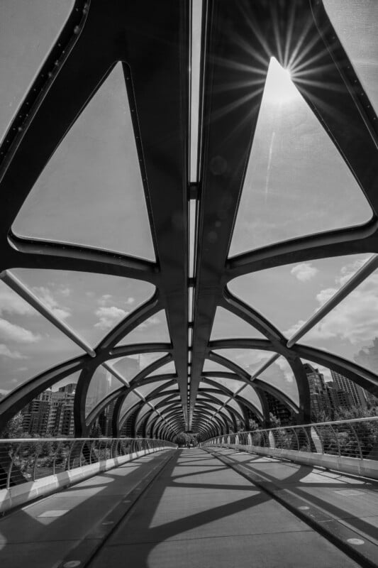 Black and white photo of a modern, geometric pedestrian bridge with sunlight streaming through a triangular section, casting shadows on the pathway below; city buildings and clouds are visible in the background.