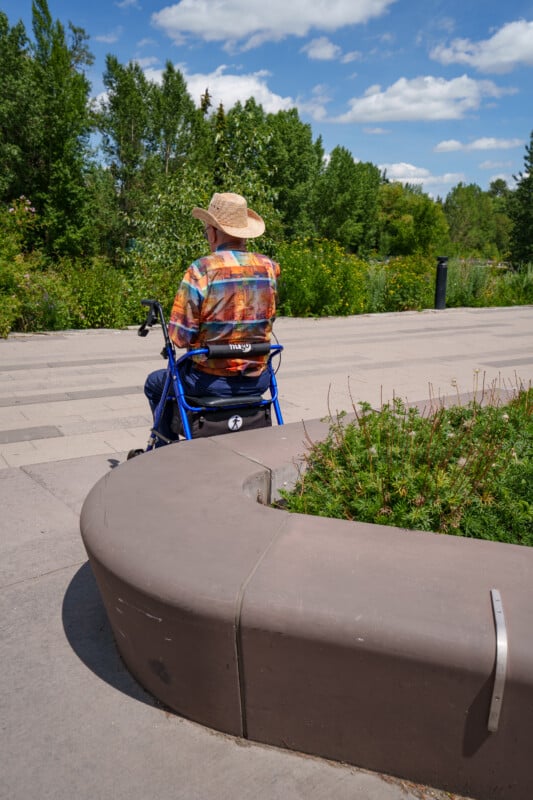 An older adult wearing a straw hat and colorful shirt sits on a blue walker at the edge of a curved bench, facing a park with green trees under a partly cloudy sky.