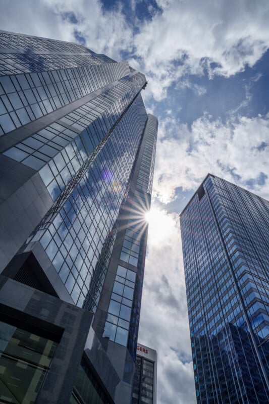 Skyscrapers with glass facades rise toward a partly cloudy sky, reflecting sunlight. The sun peeks between two buildings, creating a bright starburst effect.