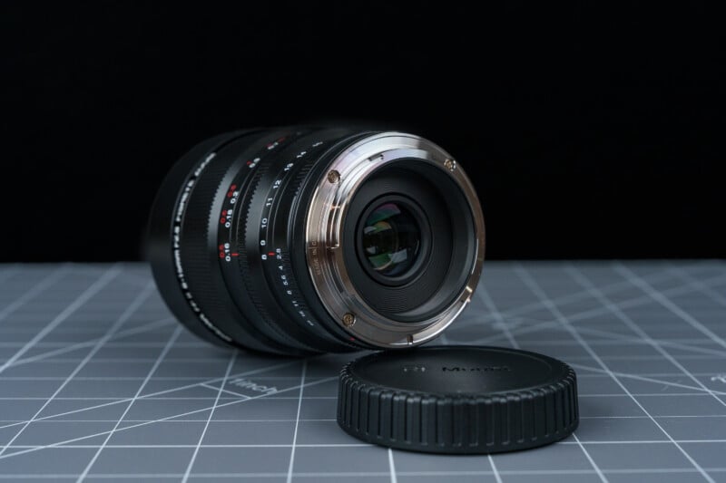 A camera lens resting on its side on a gray grid-patterned surface, with its rear glass and metal mount facing forward. A detached lens cap lies nearby. The background is black.