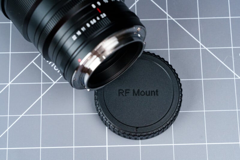 A camera lens and its detached black lens cap labeled "RF Mount" rest on a gray cutting mat with a white grid pattern.