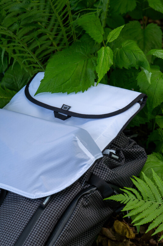 A close-up of a backpack with a white top flap and black straps, partially covered by large green leaves and surrounded by lush ferns and foliage.