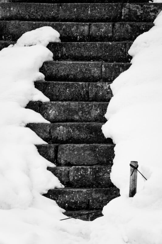 Stone steps partially covered in snow, creating a symmetrical pattern on both sides. The snow contrasts with the dark, weathered texture of the stairs, leading upwards in an outdoor setting. A wooden post with wire is visible at the bottom right.