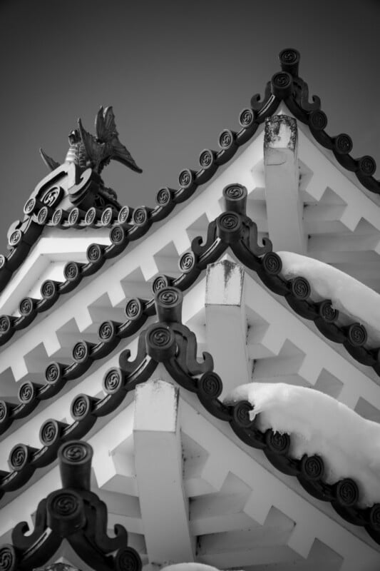 Close-up of a traditional Japanese roof with ornate, curved tiles and a decorative ornament at the peak. Snow lightly covers parts of the roof. The image is in black and white, highlighting the roof's intricate details and design.