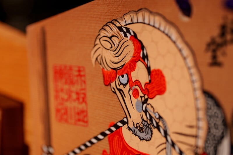 Close-up of a decorative illustration showing a stylized horse with intricate red and black patterns. The background includes a hexagonal pattern and faint calligraphy. Colors are vivid, emphasizing the horse's detailed features.